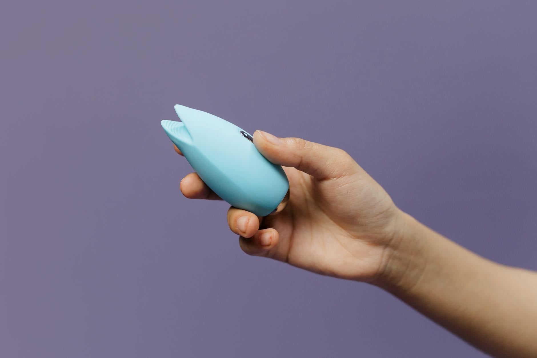 A blue silicone clitoral vibrator held in hand against a purple background, symbolizing self-care and intimate reconnection